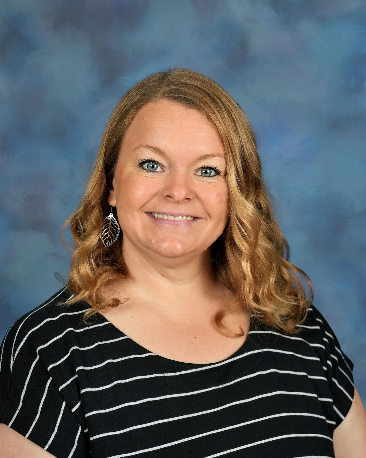 Smiling woman with wavy blonde hair wearing striped shirt and a feather earring against a blue textured background. Casual and friendly tone.